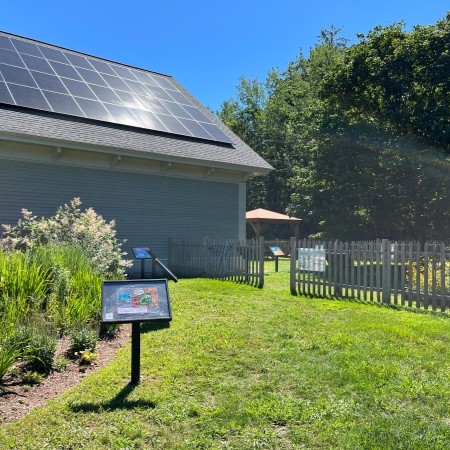 A sunny view of the Story Garden, featuring a fenced reading area with outdoor story panels. The library building, with solar panels on its roof, is visible in the background.