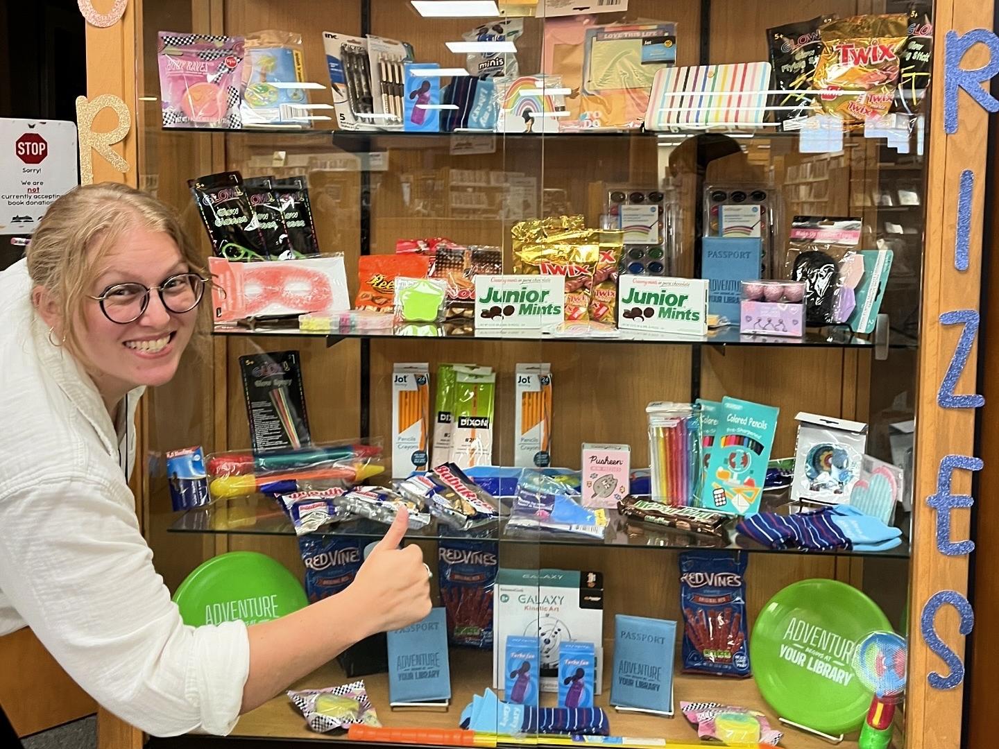 Molly smiling and giving a thumbs up at the Teen Prizes cabinet for summer reading. 